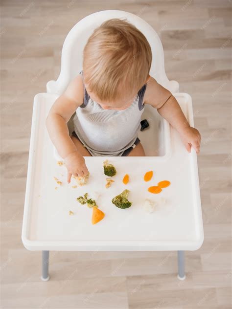 Bebé sentado en una trona, explorando trozos de verduras de colores en un plato.
