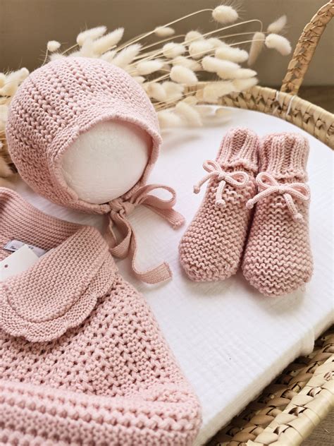 Imagen de conjunto de capota y patucos de bebé modelo Ángel, tejidos a mano en tono rosa empolvado sobre fondo rústico con flores decorativas