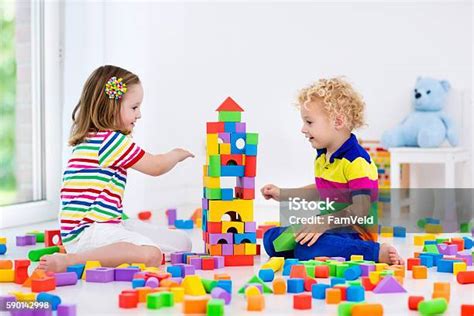 Dos niñas riendo y construyendo una torre de bloques durante una cita de juego, con una figura adulta en el fondo observando