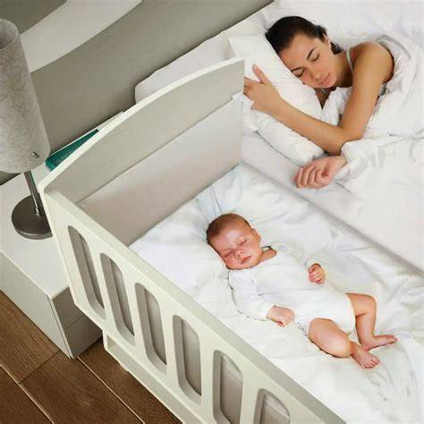 Bebé durmiendo plácidamente en una cuna colecho junto a la cama de sus padres, con los padres observándolo